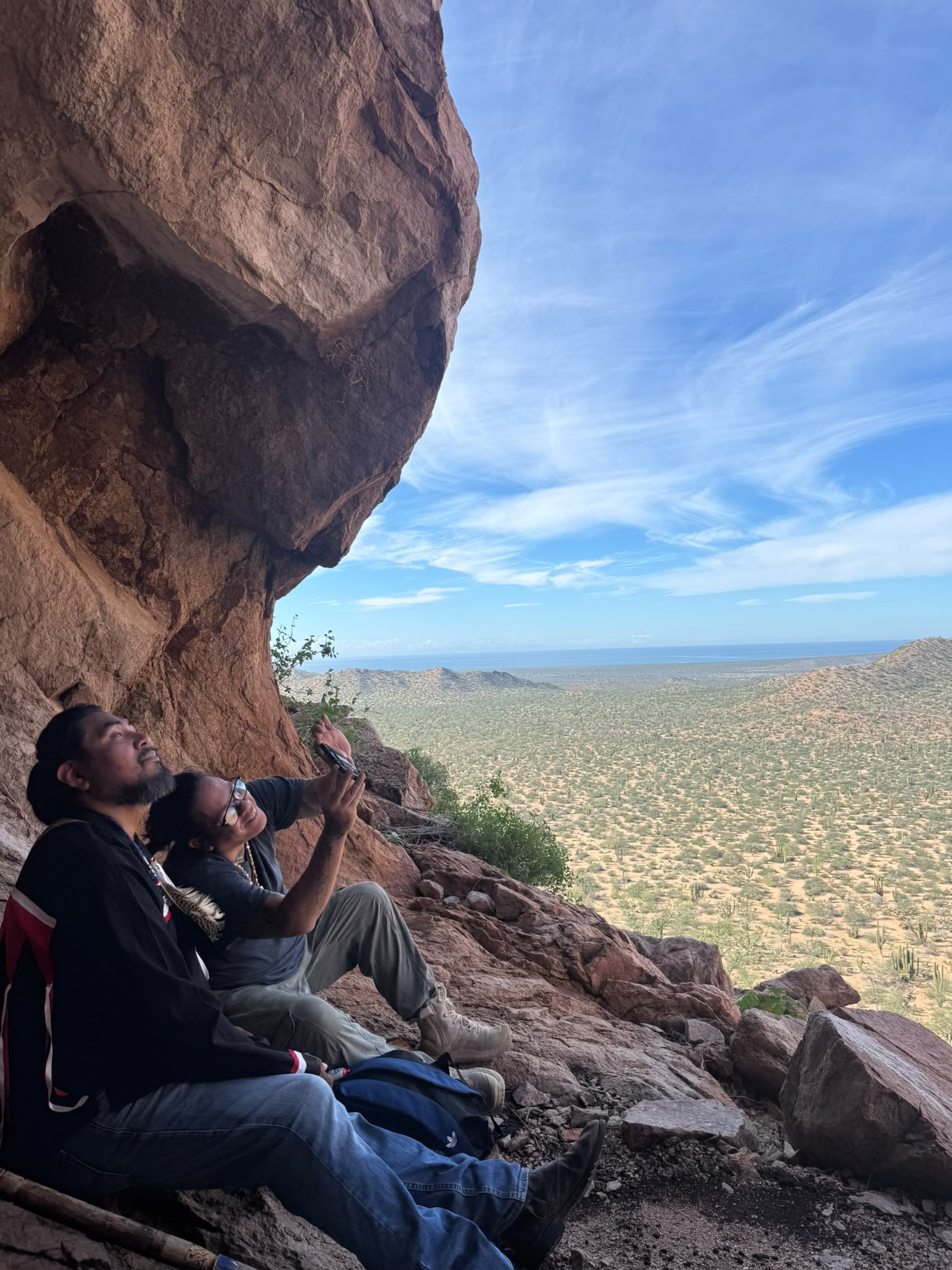 Two seekers resting beneath the cave overhang, overlooking the Sonoran desert and the Sea of Cortez